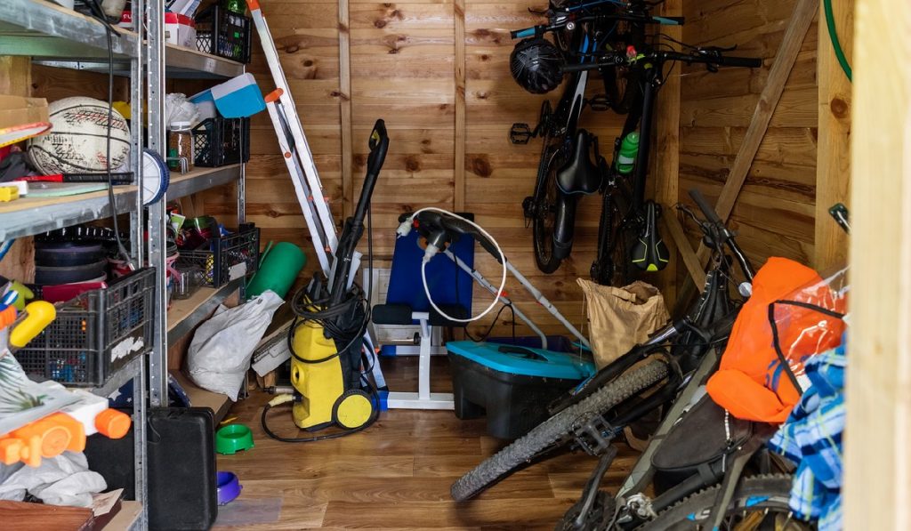 Suburban home wooden storage utility unit shed with miscellaneous stuff on shelves, bikes, exercise machine, ladder, garden tools and equipment. Messy and chaos at house yard barn.