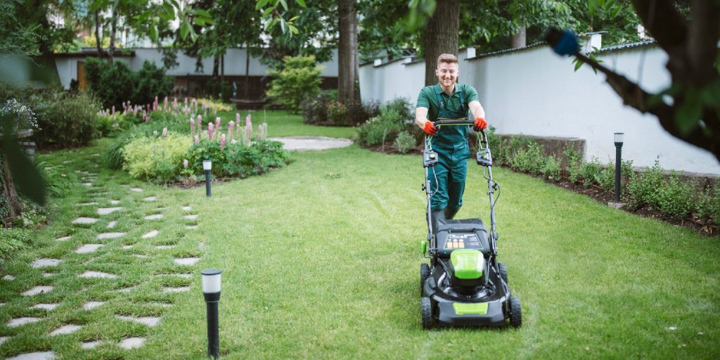 Full length gardener mows the lawn with a mower in beautiful garden.